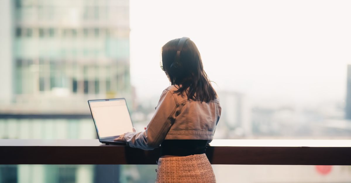 A woman wearing headphones works on her laptop overlooking a cityscape from an office or apartment balcony.